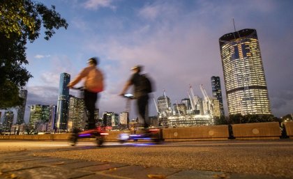 two people on scooters riding along a footpath at dusk. The city skyline is in the background